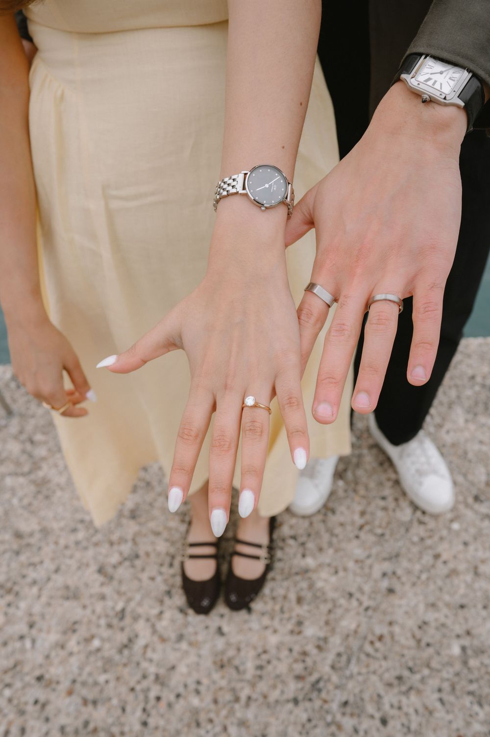 Newlyweds displaying wedding rings and watches on outstretched hands, bride wearing white nail polish and diamond engagement ring.
