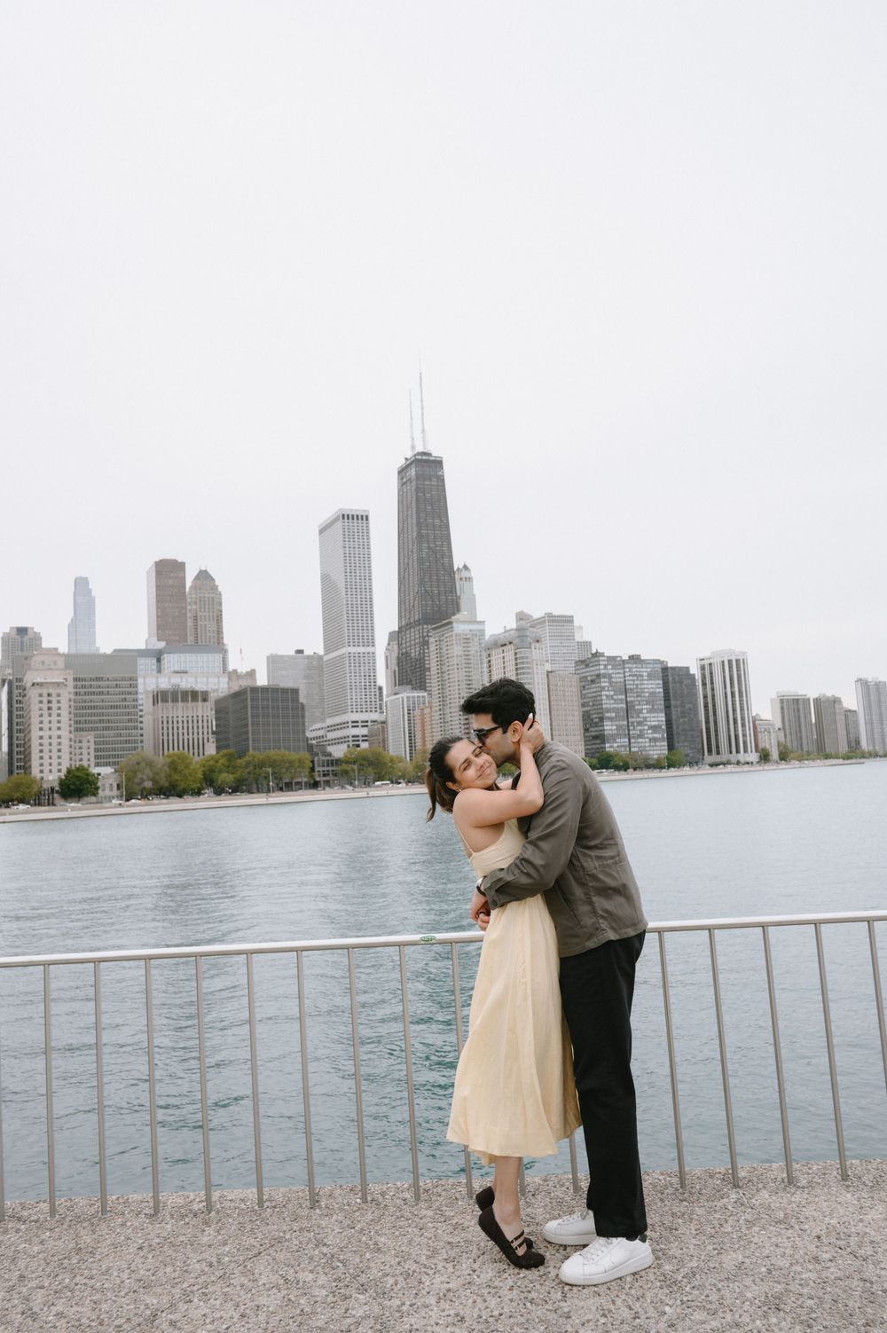 Couple embracing and kissing by Lake Michigan with Chicago skyline in background on overcast day.