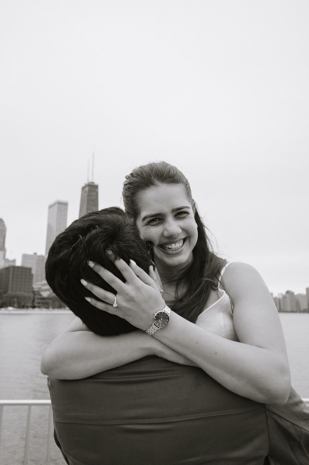 Black and white photo of a smiling woman hugging a man with a city skyline in the background.