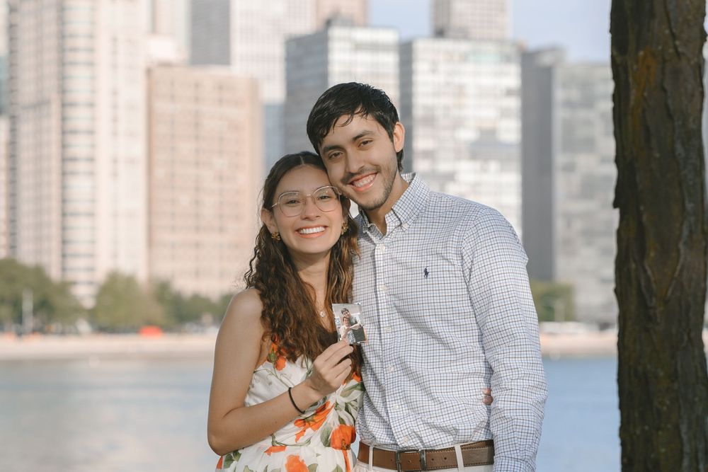 A smiling couple poses near a waterfront with a city skyline backdrop on a sunny day.