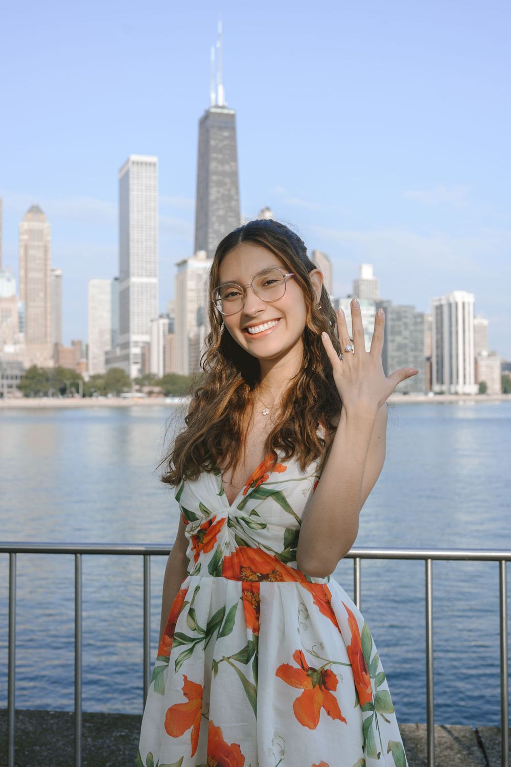 Smiling young woman in floral dress waving with Chicago skyline and Lake Michigan in the background.