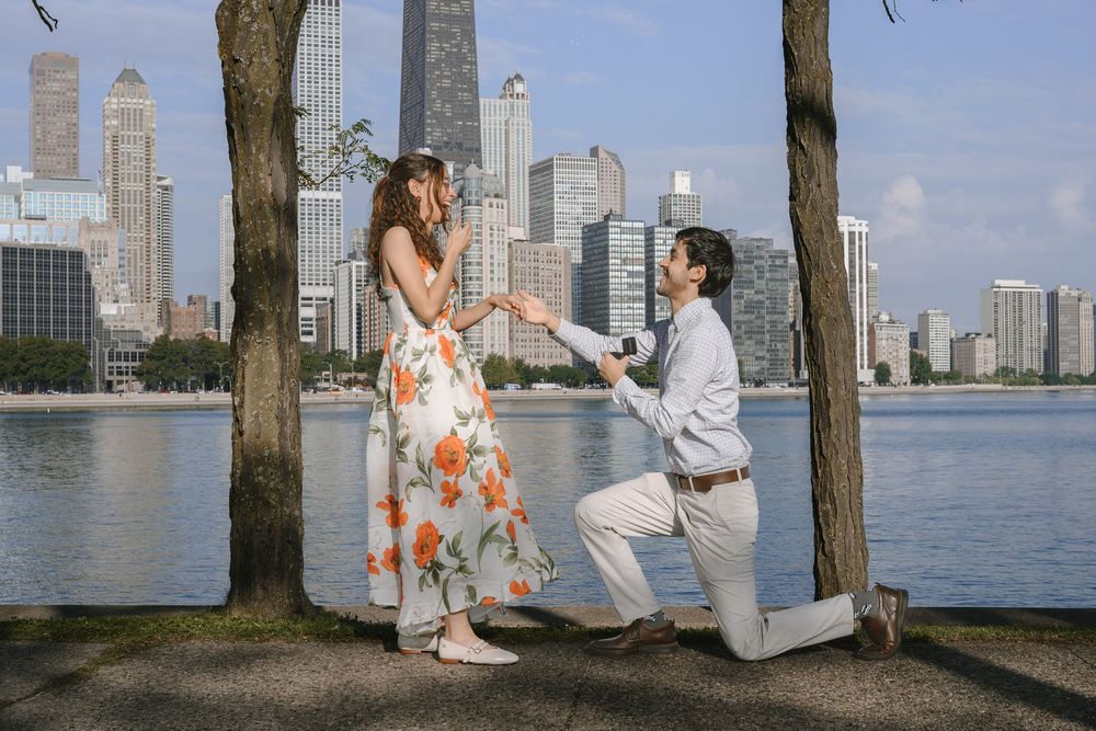 Man proposes to woman on one knee by waterfront with Chicago skyline in background, woman in floral dress reacts joyfully.