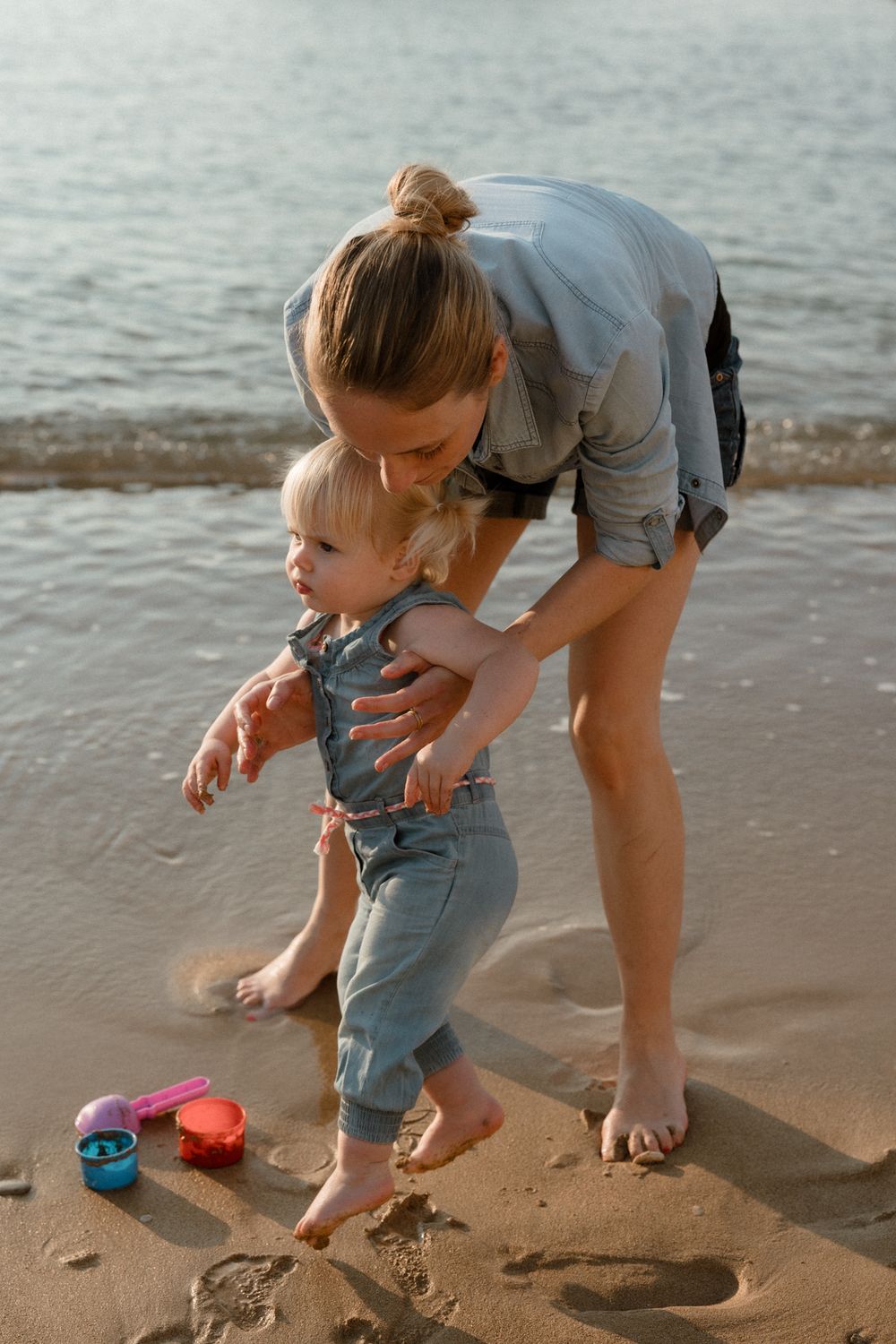 A mother helps her toddler walk on a sandy beach near the water, with colorful toys nearby on a sunny day.