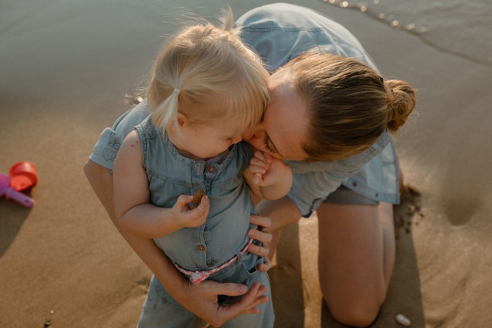 Mother kissing toddler girl in denim dress on sandy beach during golden hour.
