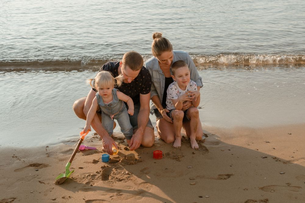 Family with two young children playing with colorful sand toys on a sunny beach near calm ocean waves.