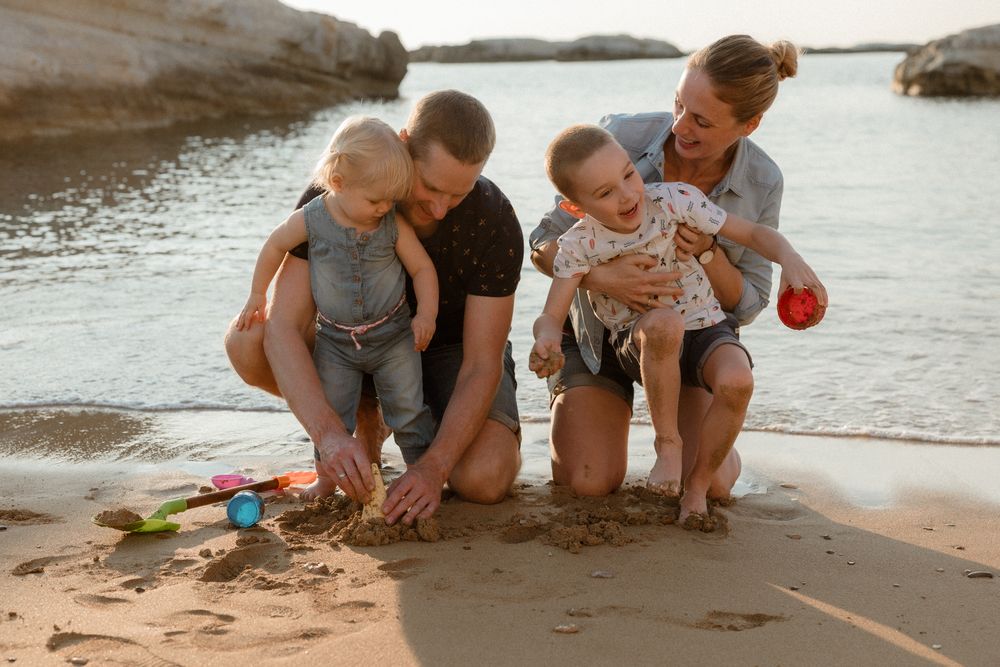 Family of four playing on sandy beach at sunset, parents and two toddlers enjoying sand toys by calm water.