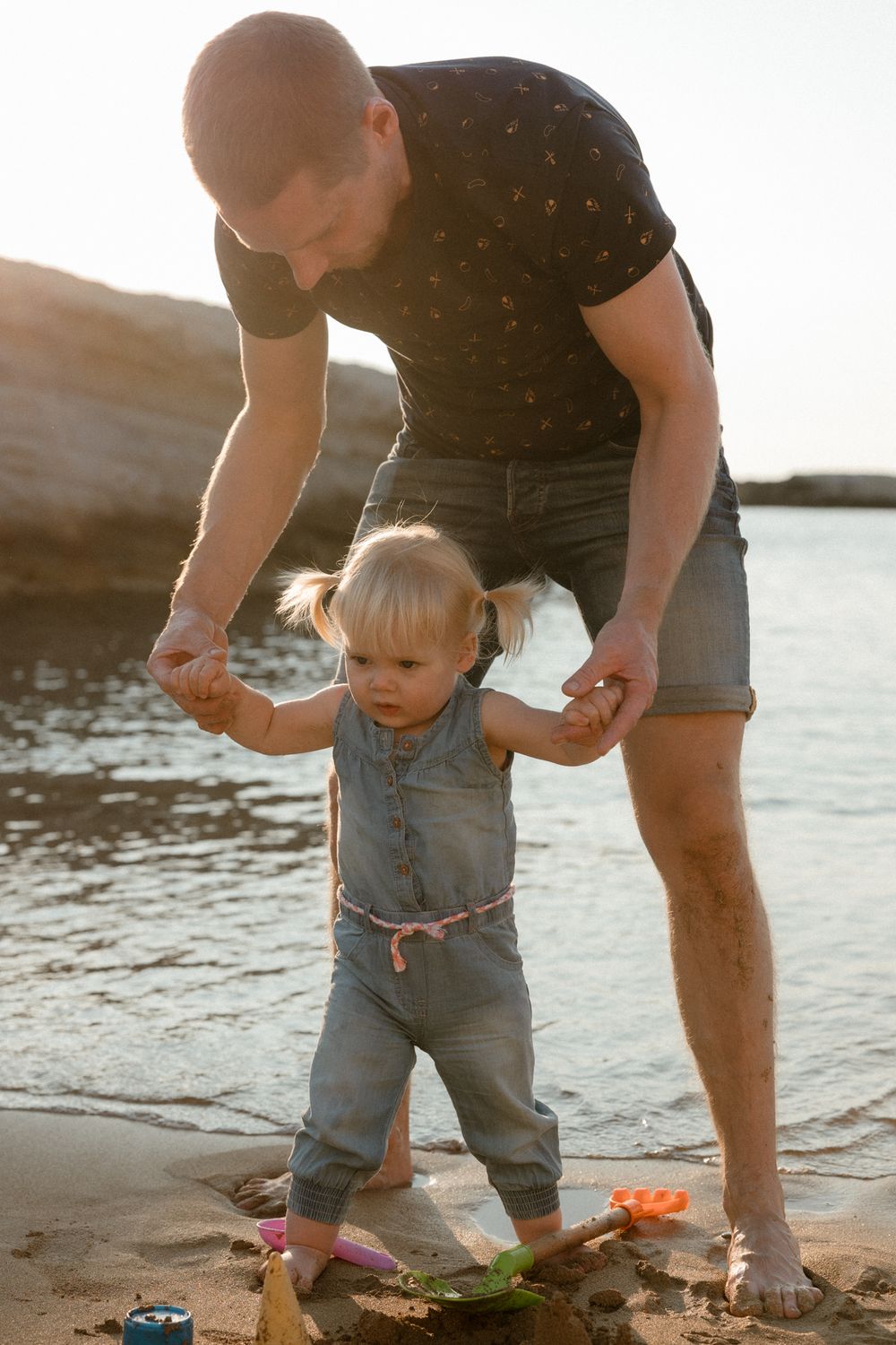 Father helps toddler girl walk on sandy beach near water during golden hour sunset.