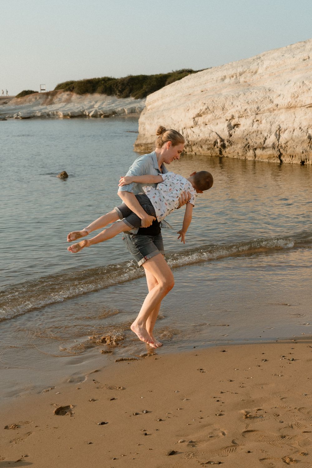 A woman playfully lifts a child on a sandy beach near calm waters and white rocky cliffs at sunset.