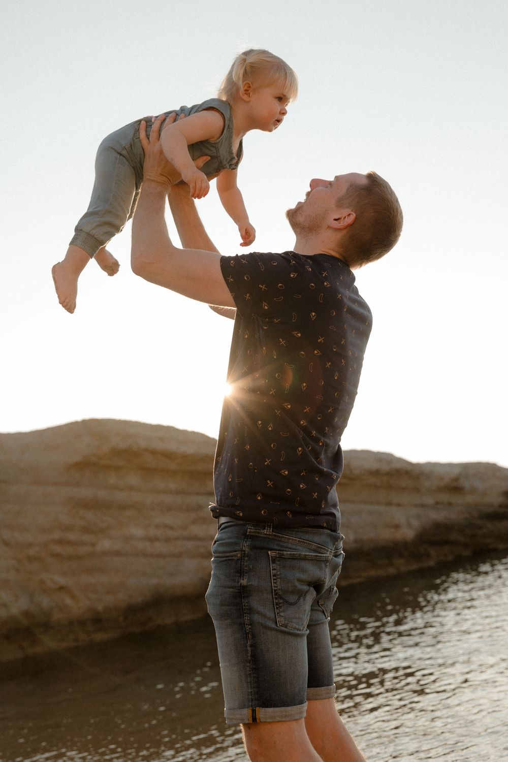 Father lifting toddler in the air at sunset by rocky coastline, sharing a joyful moment outdoors.