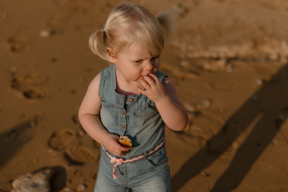 Blonde toddler girl in denim romper eating a snack on a sandy beach in warm golden sunlight.