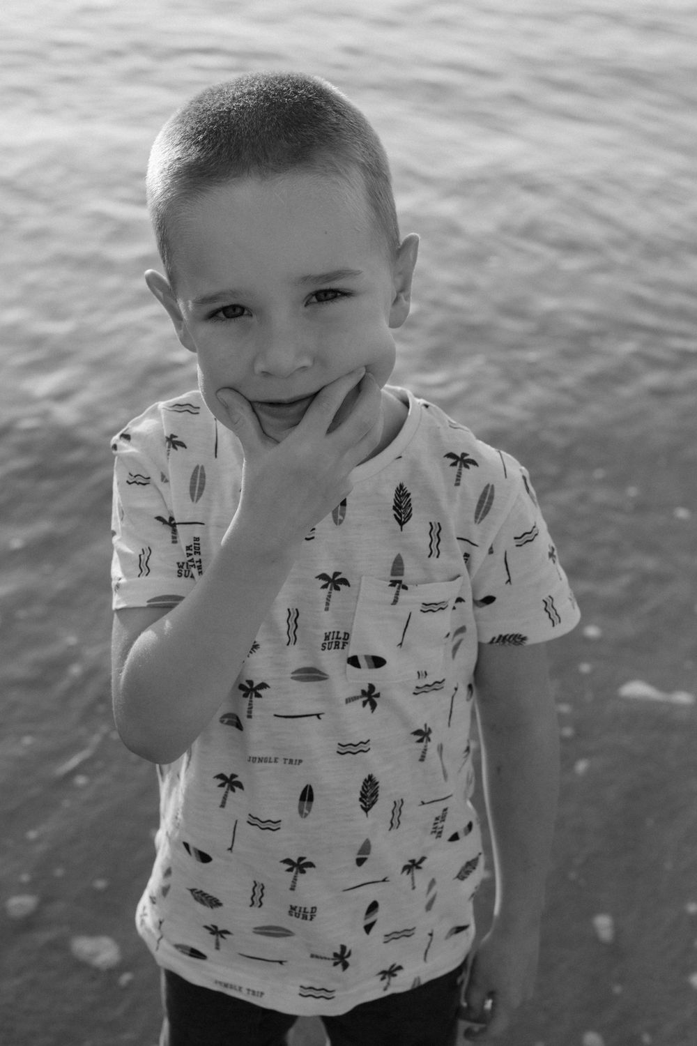 Black and white photo of a smiling young boy in a printed t-shirt posing by the water's edge.