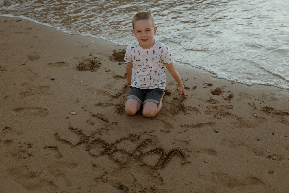 Young boy sitting on sandy beach near ocean waves with "LEON" written in the sand beside him.