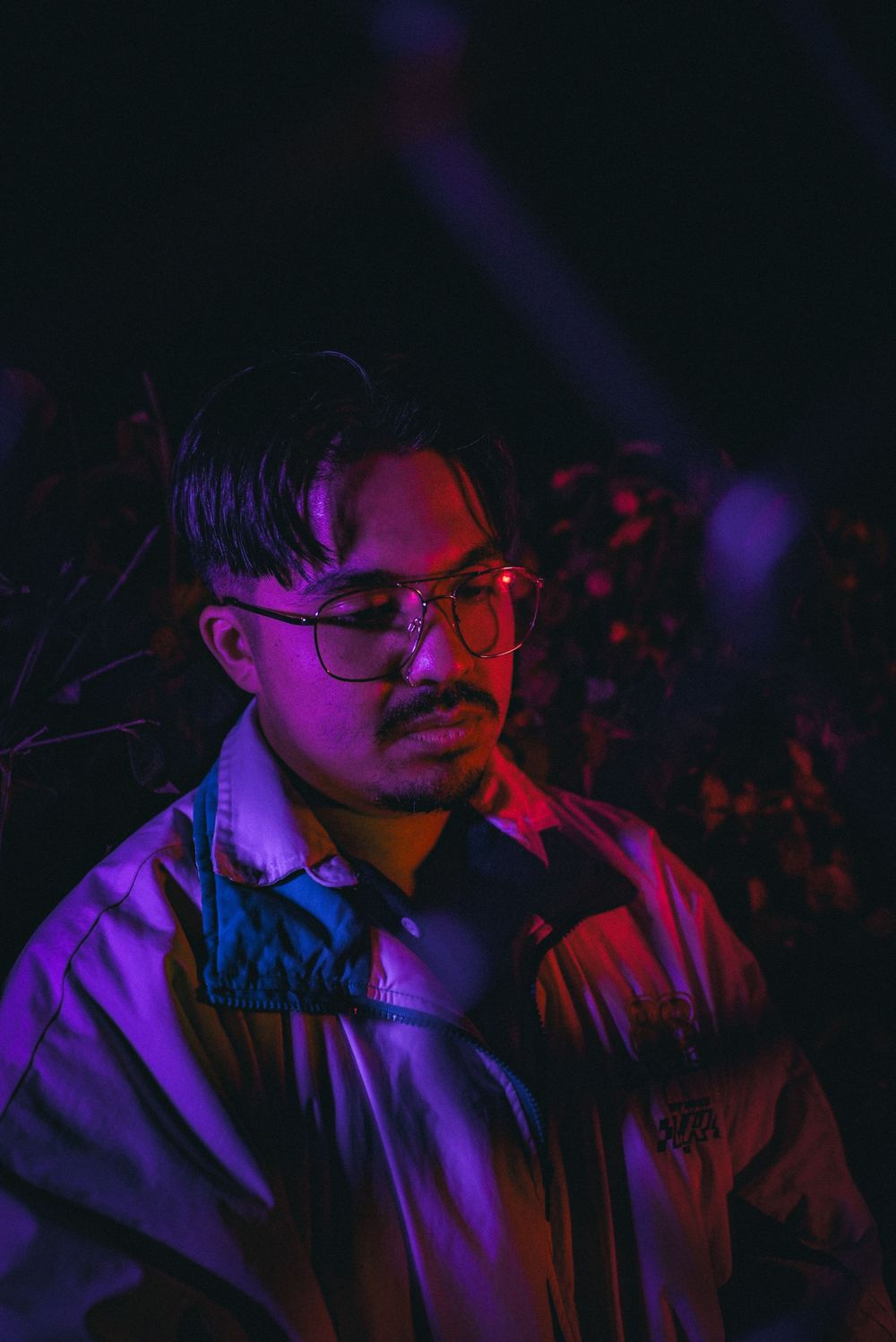 Young man with glasses and mustache illuminated by neon purple and red lights at night outdoors.