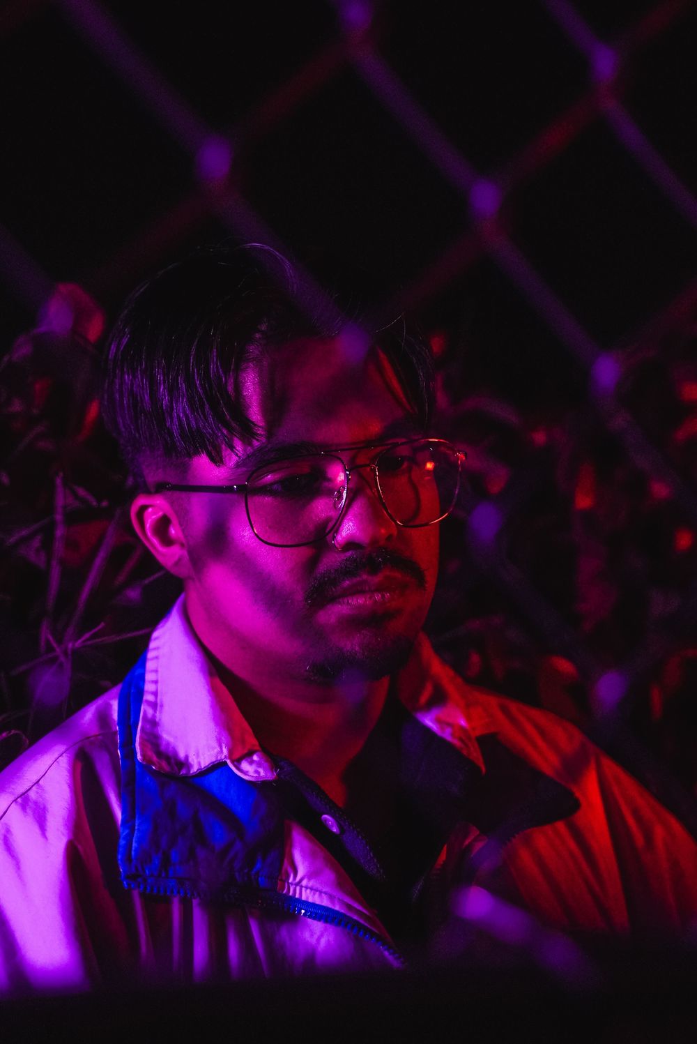 Man with aviator glasses and mustache posed against chain-link fence under dramatic purple and blue neon lighting at night.