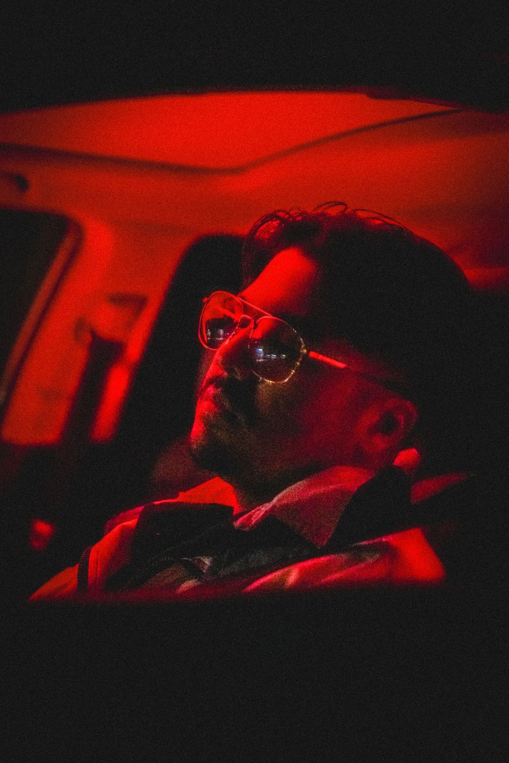 Man with aviator glasses bathed in red light inside a dark car, creating a moody cinematic atmosphere.