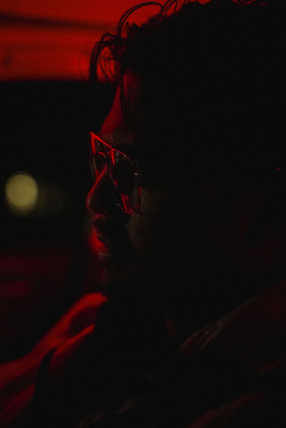 A man with glasses illuminated by dramatic red neon light in a dark moody portrait shot.