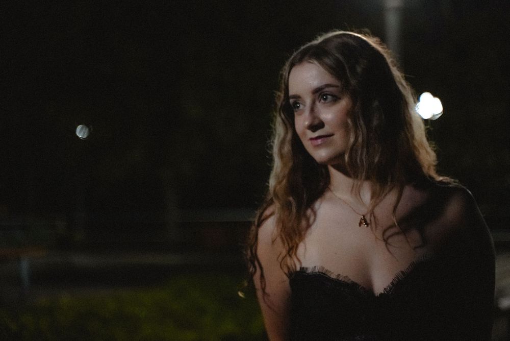 A young woman with curly hair in a black dress poses outdoors at night under soft lighting.