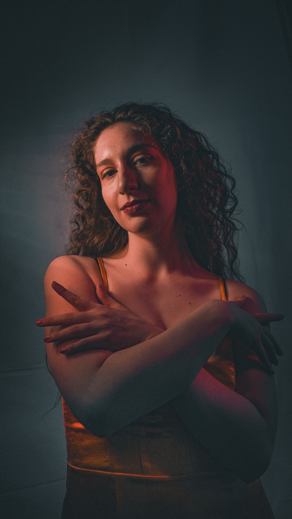 A curly-haired woman in an orange top crosses her arms in a dramatic warm-toned studio portrait.
