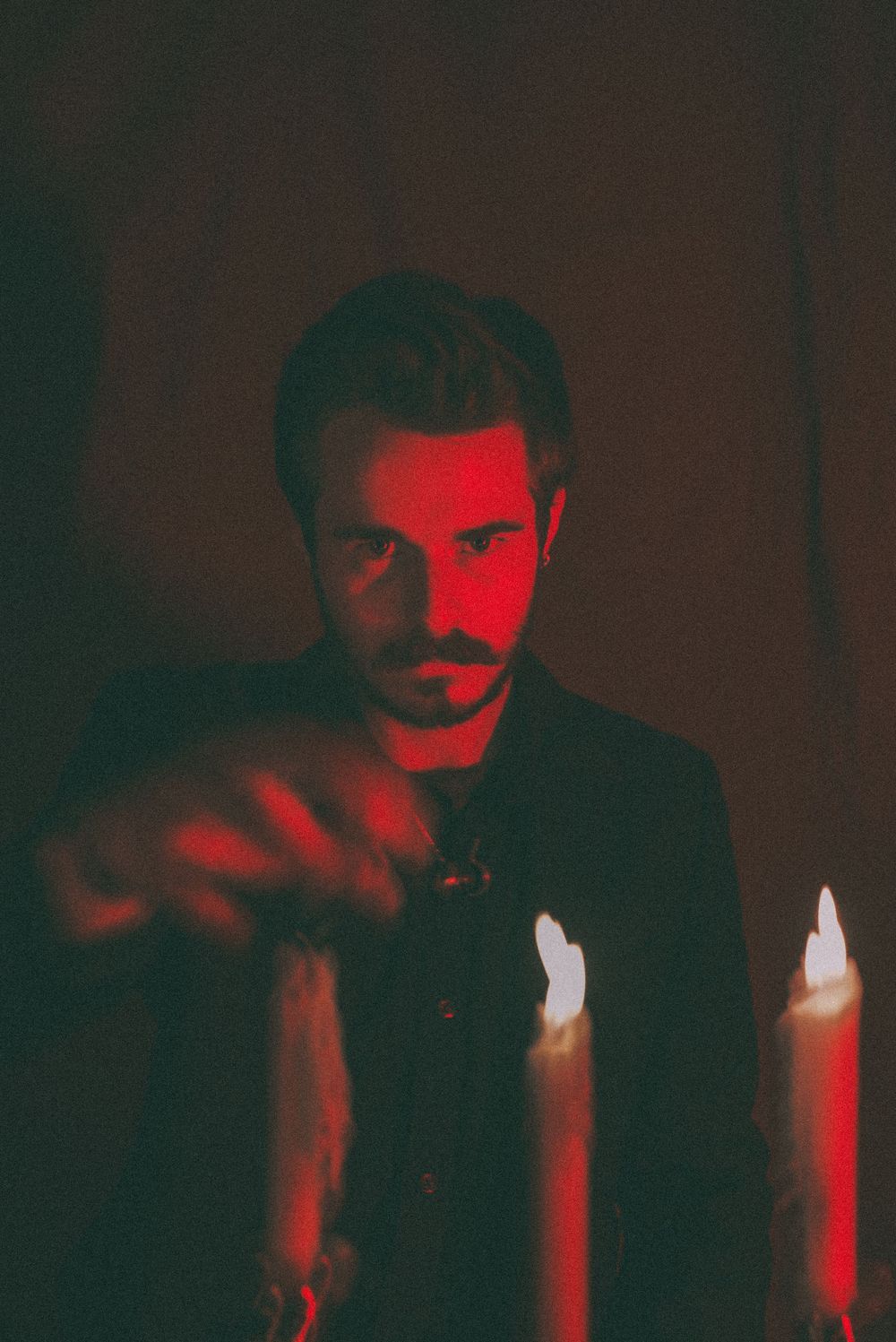 Man with beard in dark red candlelit room reaching toward camera with moody atmospheric lighting.