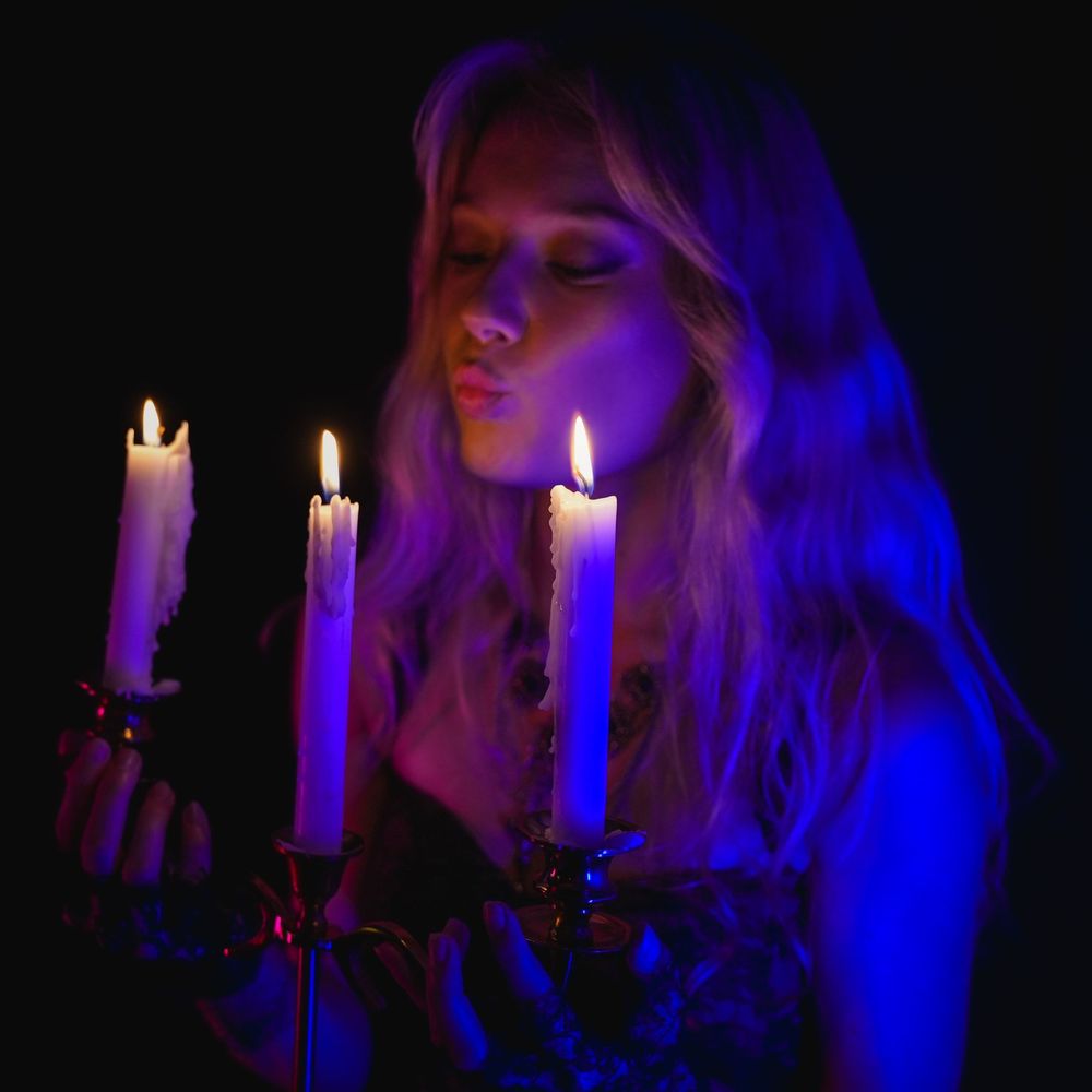 Blonde woman blowing out candles on ornate candelabra bathed in blue and purple light.