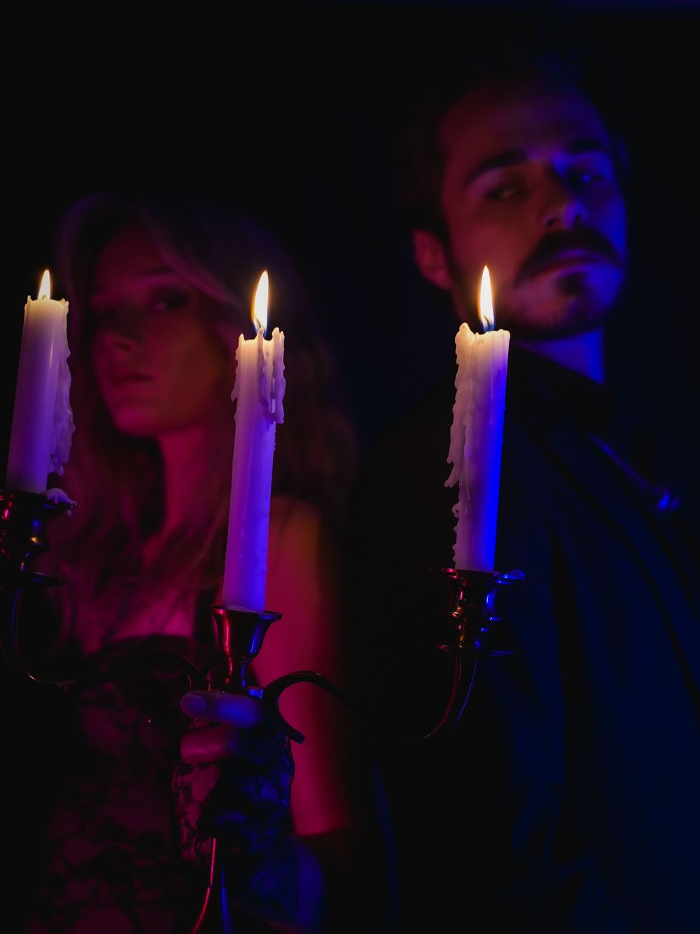 Two people holding lit candles in candlesticks in a dark, moody, gothic-style setting with blue and warm lighting.
