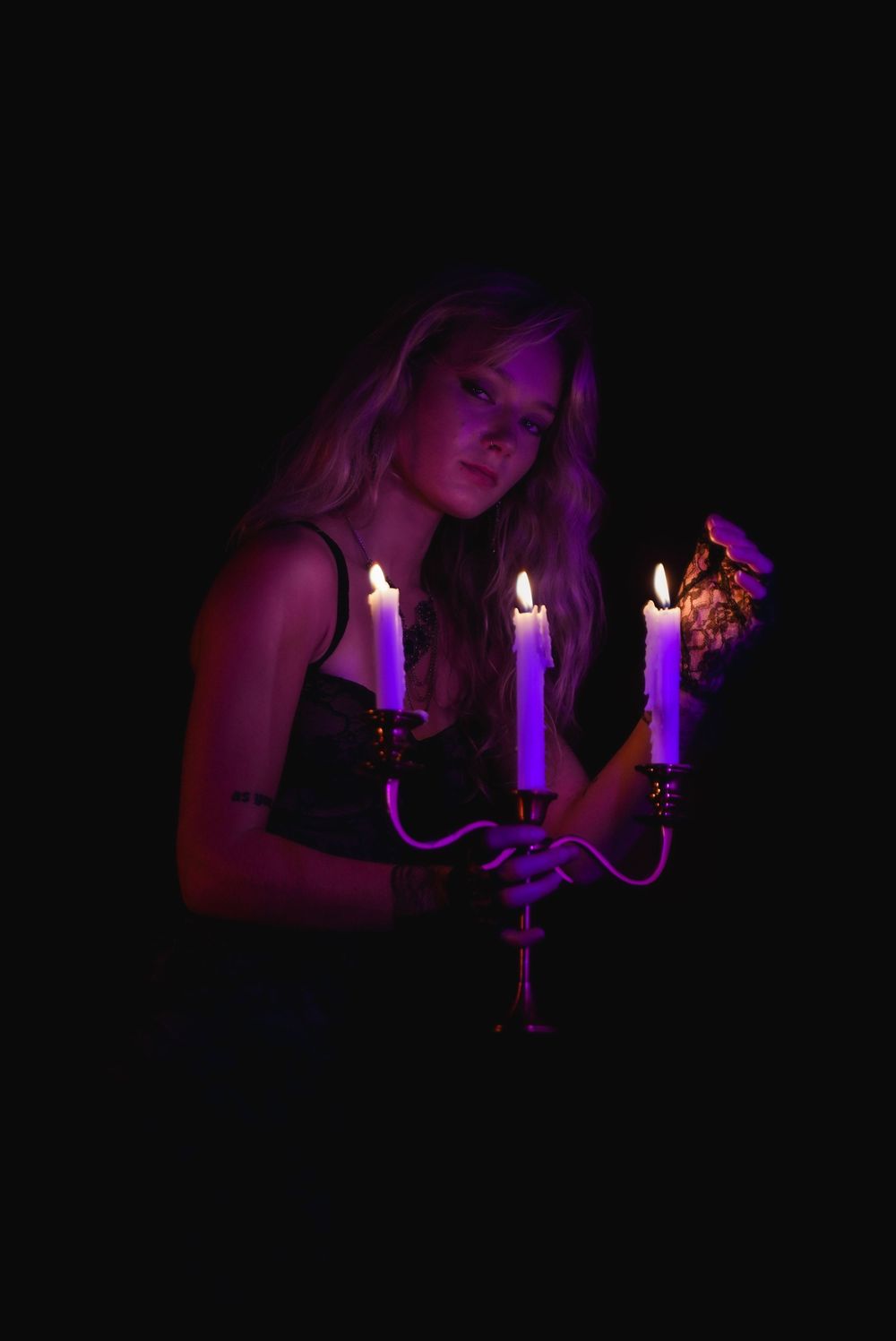 A young woman holds a candelabra with three glowing purple candles in a dark, moody setting.