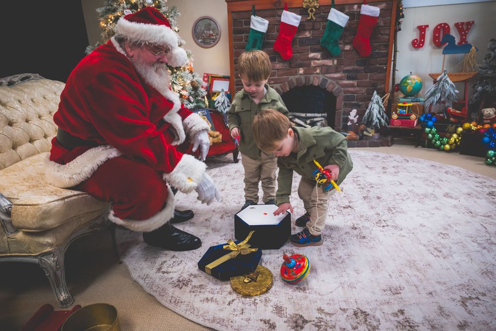 Two children in elf costumes play with toys beside Santa Claus in a festive Christmas-decorated living room.