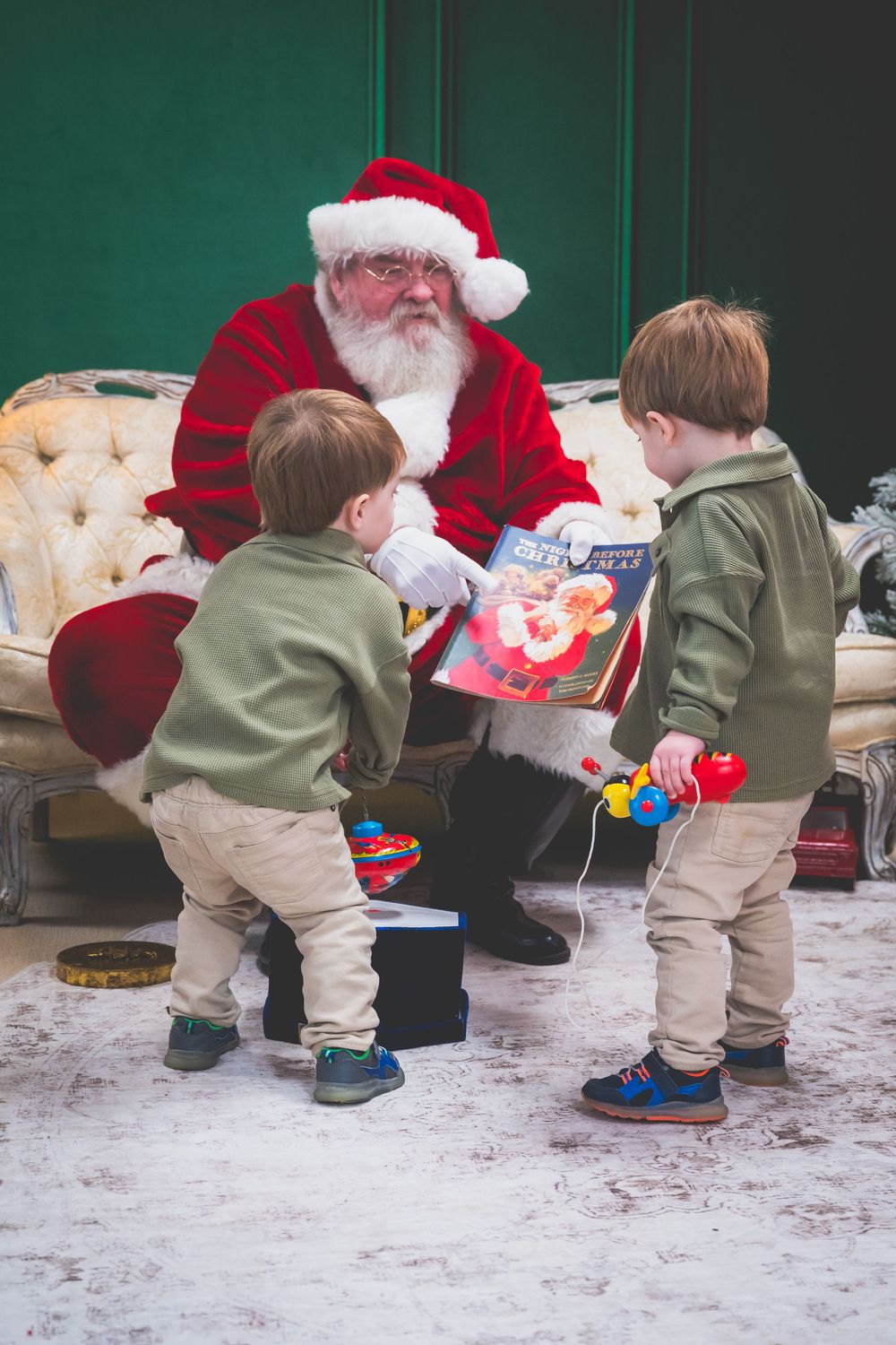 Santa Claus reads a book to two young boys in green sweaters during a festive Christmas visit.