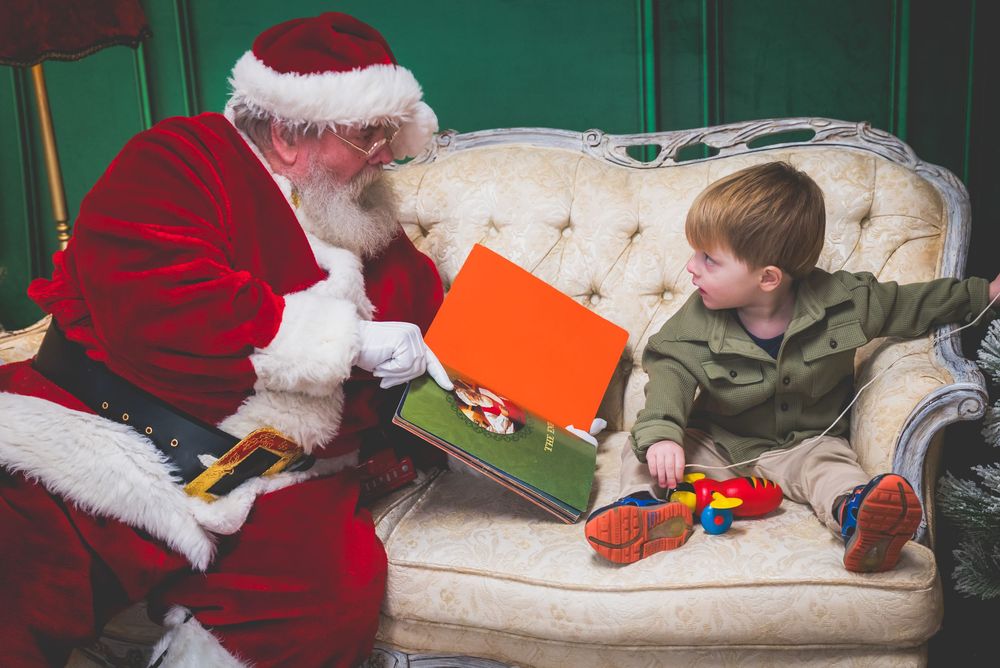 Santa Claus showing a colorful book to a young red-haired boy sitting on an elegant white sofa during Christmas.