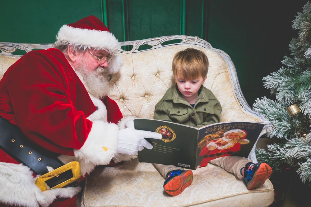 Santa Claus reads a festive Christmas book with a young child on a decorated holiday sofa near a Christmas tree.
