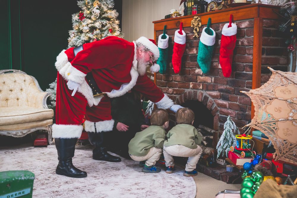 Santa Claus kneels by a decorated fireplace with stockings alongside two small children opening gifts.