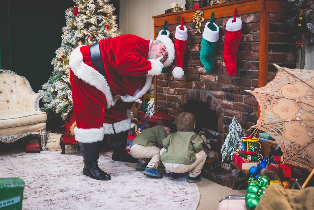 Santa Claus bending down near a brick fireplace with Christmas stockings, gifts, and a decorated tree in a cozy holiday scene.