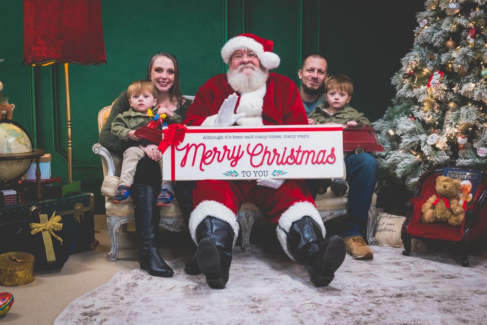 Family posing with Santa Claus holding a Merry Christmas sign beside a decorated Christmas tree.