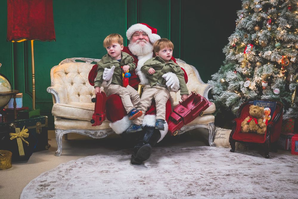 Two children sitting with Santa Claus on a cream sofa beside a decorated Christmas tree in a festive holiday setting.