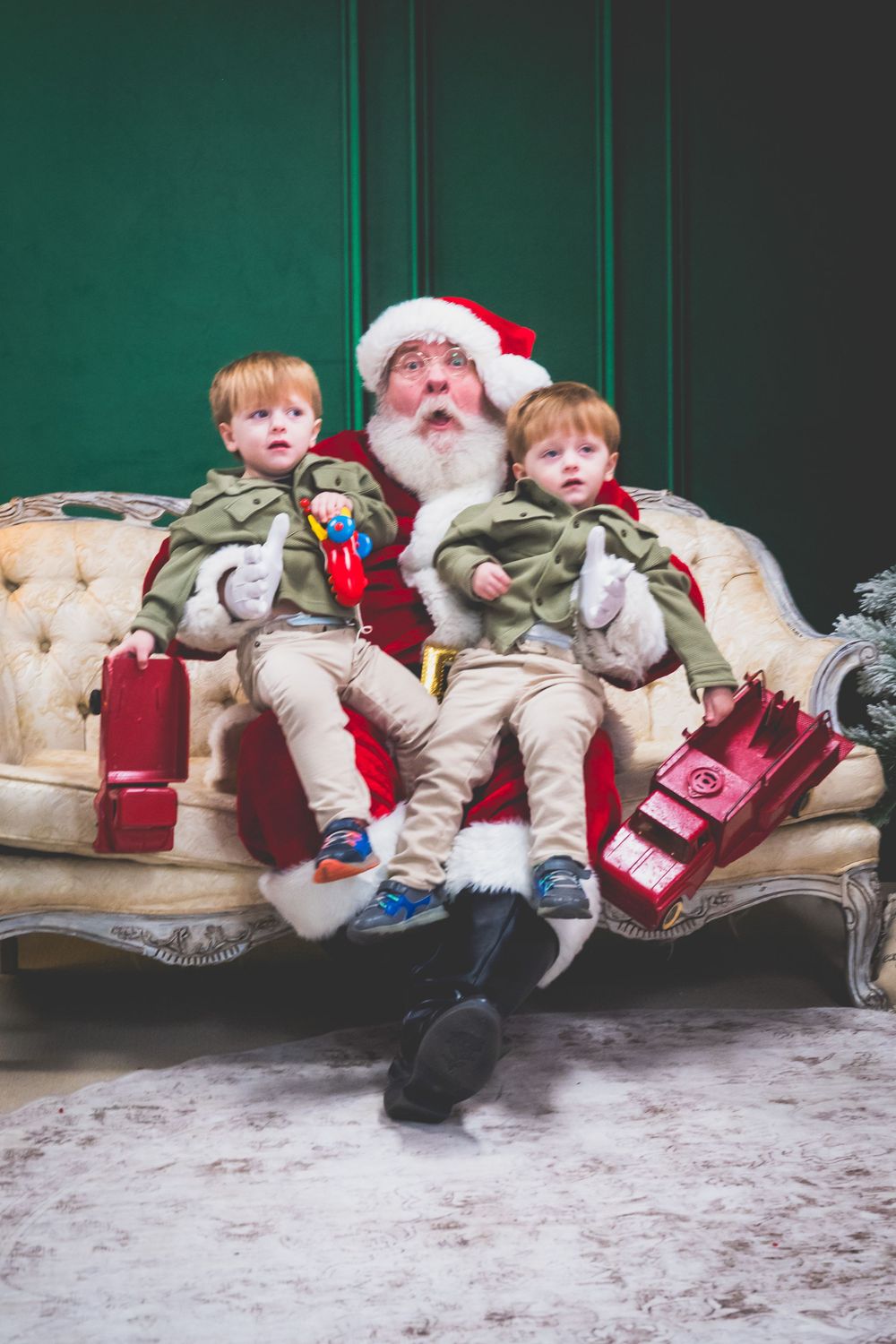 Two toddlers sitting on Santa Claus's lap on a vintage sofa, holding red toy trucks during a Christmas photo session.