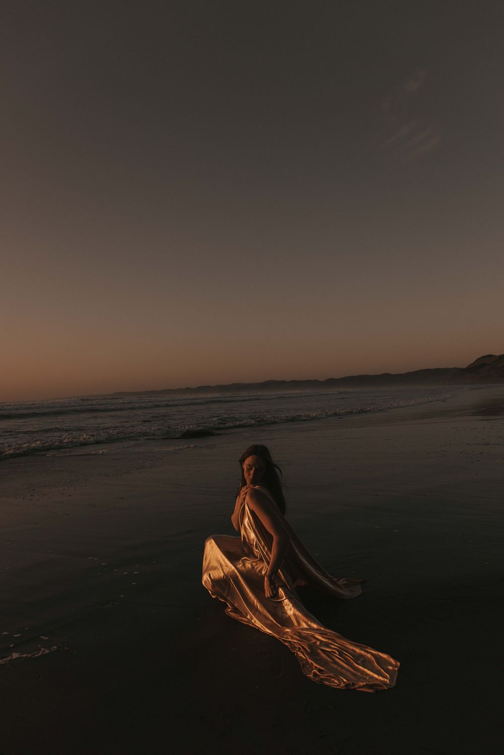 Woman in flowing golden gown poses gracefully on a dark beach at sunset with ocean waves and twilight sky.