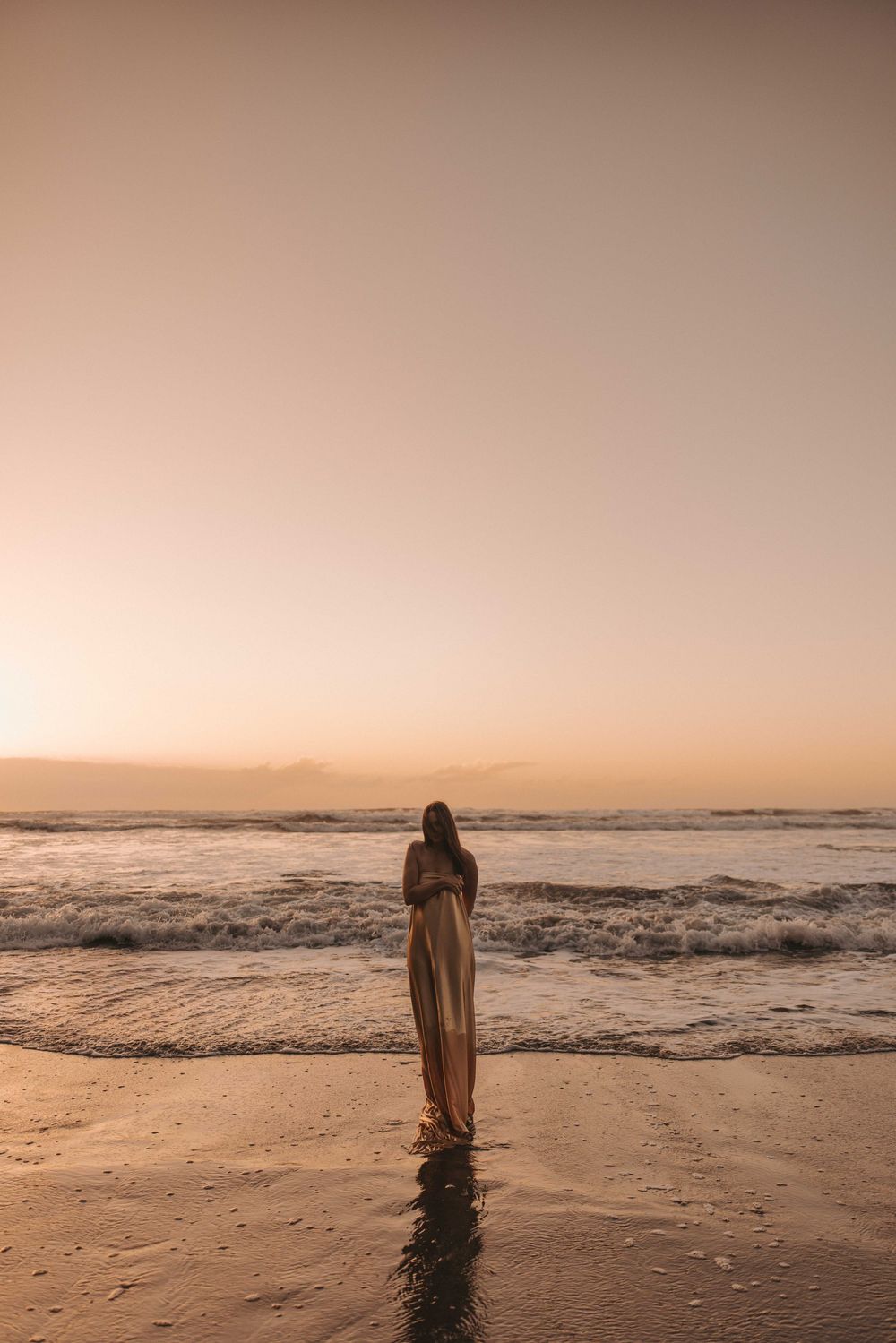 Woman in flowing dress standing on a sandy beach at golden sunset with ocean waves.