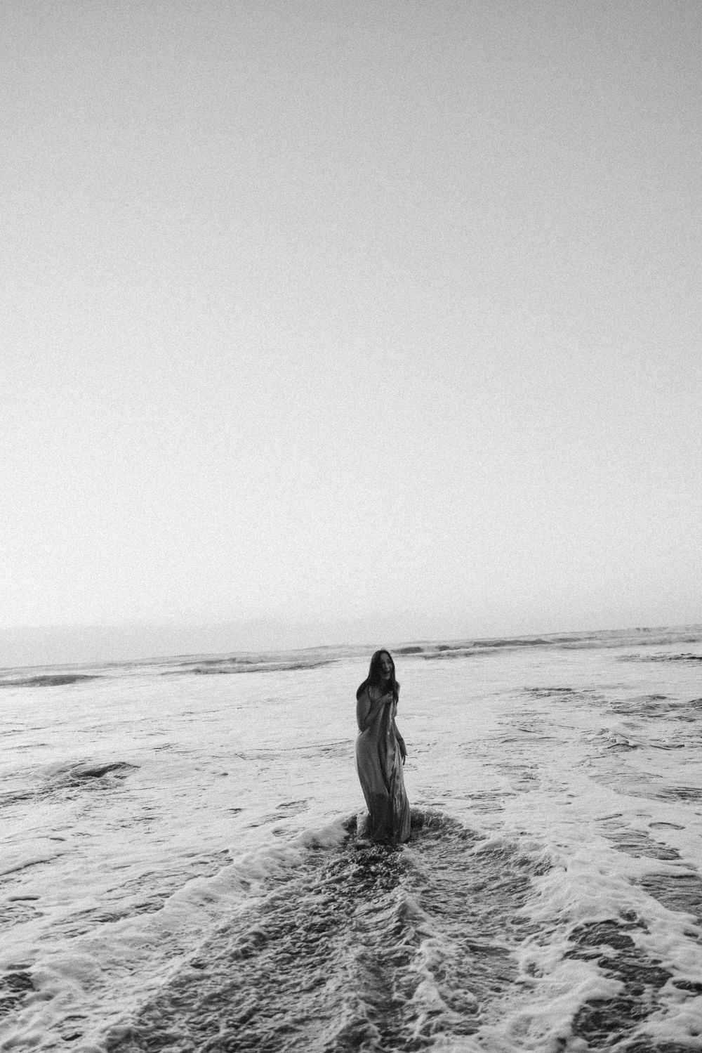 Black and white photo of a woman in a flowing dress standing in ocean waves on a beach.