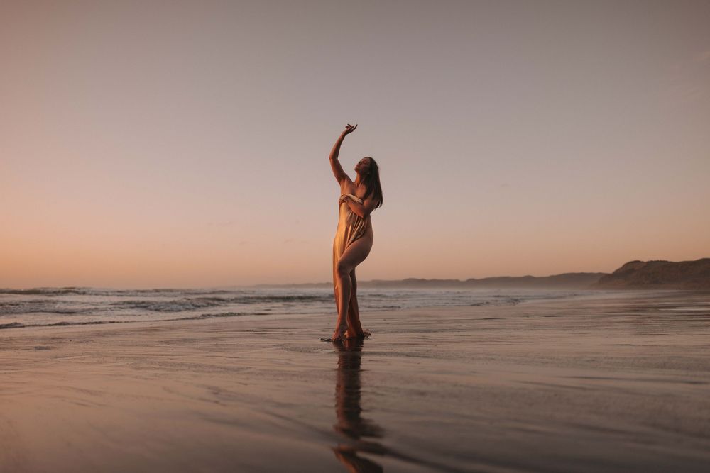 A woman in a flowing dress poses gracefully on a wet beach at sunset with waves and golden sky.