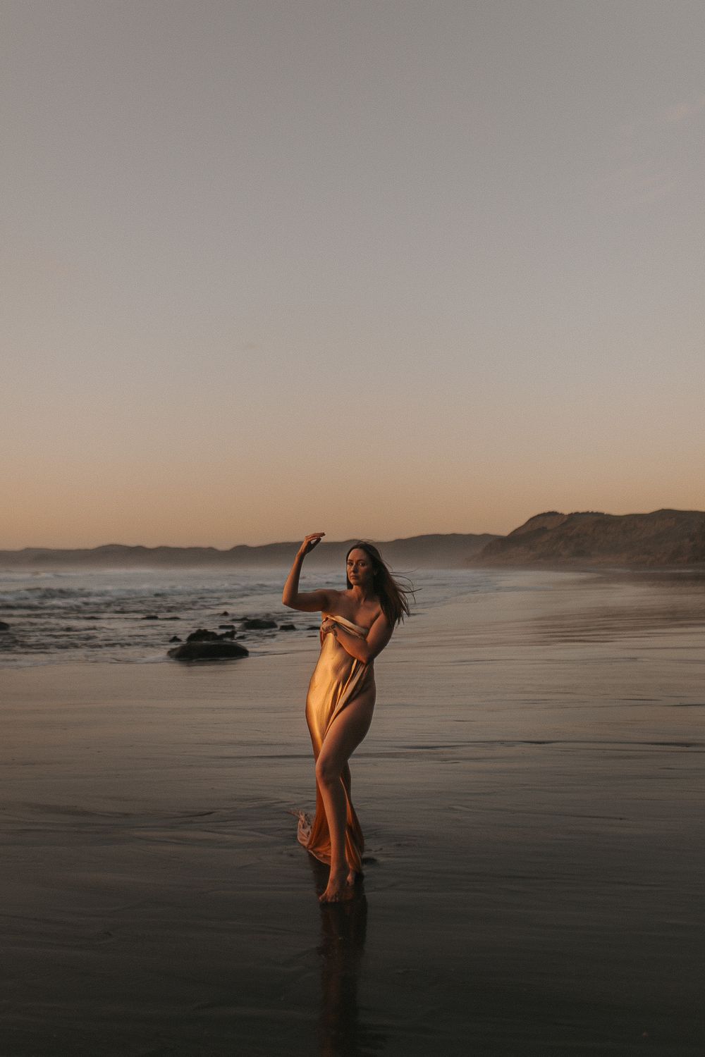 Woman in flowing dress standing in ocean waves at sunset with mountains in background.