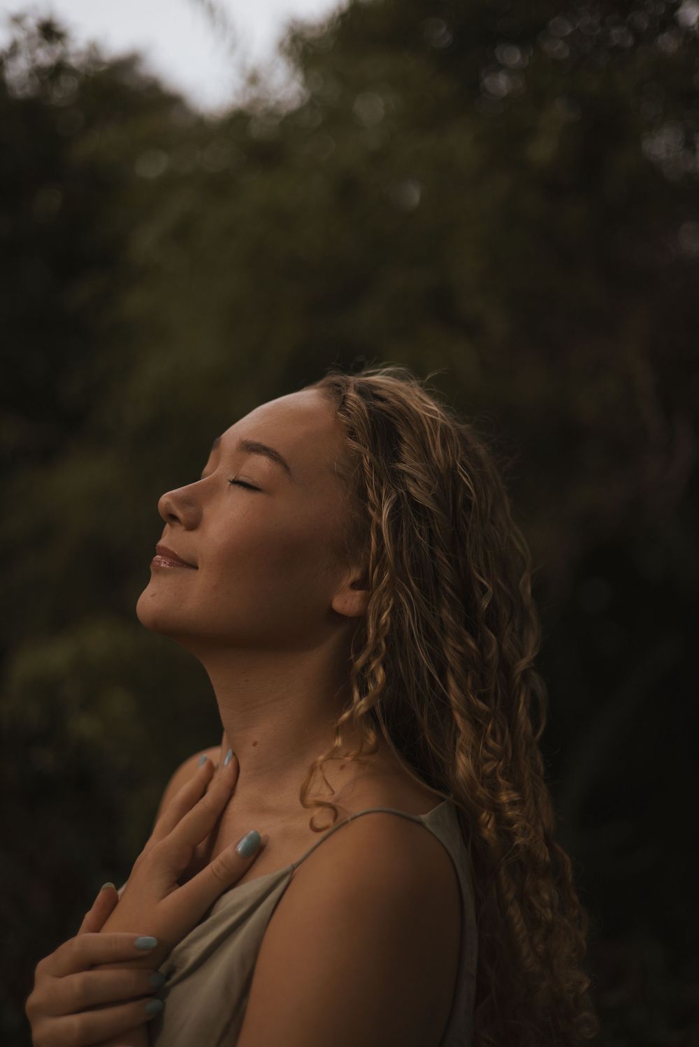 Young woman with curly blonde hair, eyes closed, smiling peacefully outdoors with lush green trees in background.