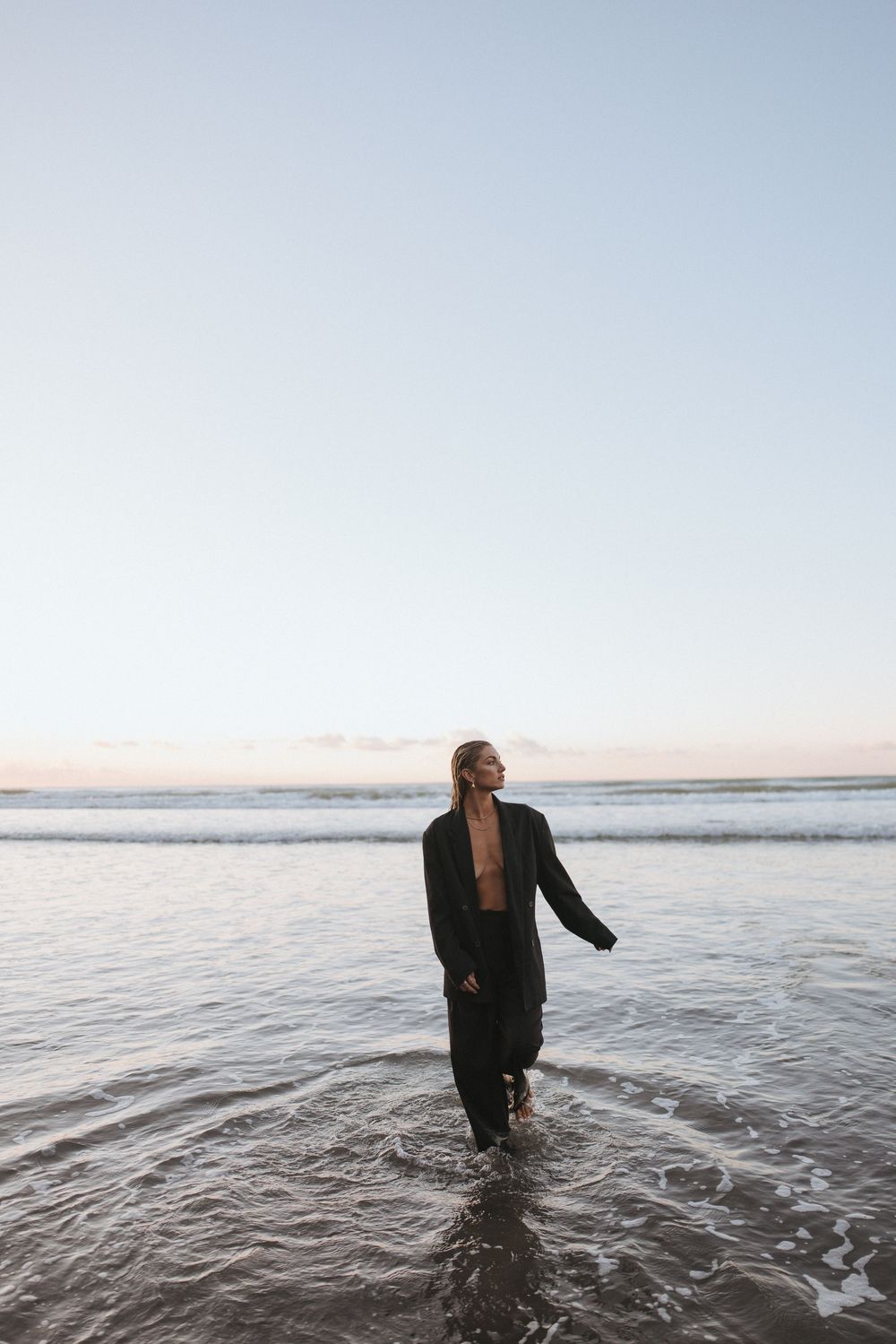 Woman in black open jacket wading in ocean waves at dusk on a serene beach.