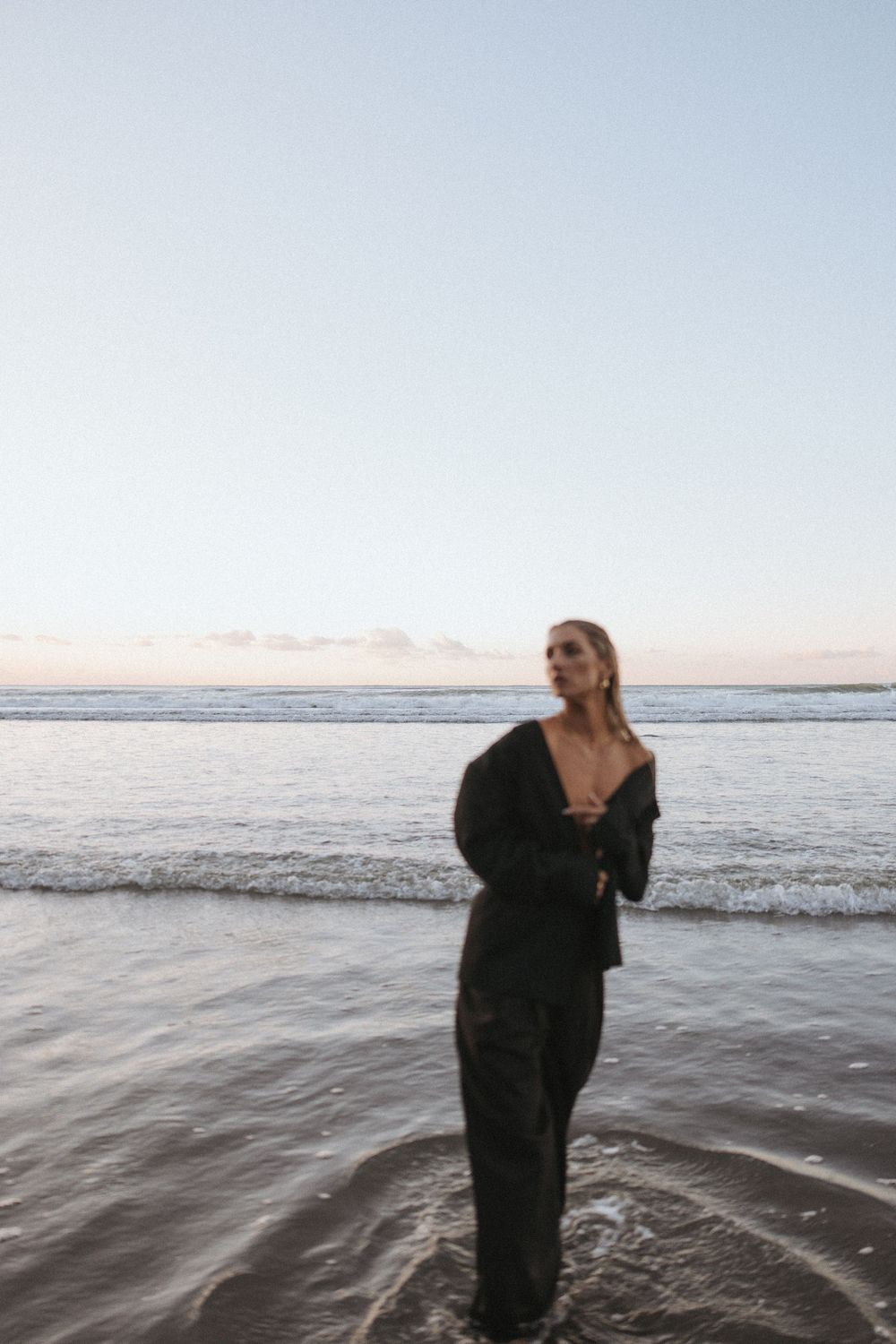 Woman in black outfit standing in shallow ocean waves at dusk on a beach.