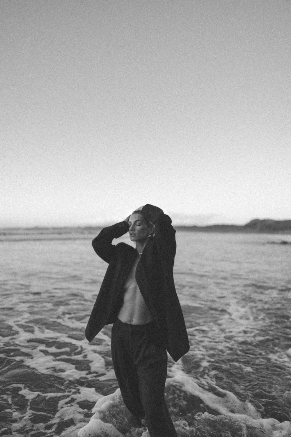 Black and white photo of a person standing in ocean waves wearing an open jacket on a beach at dusk.