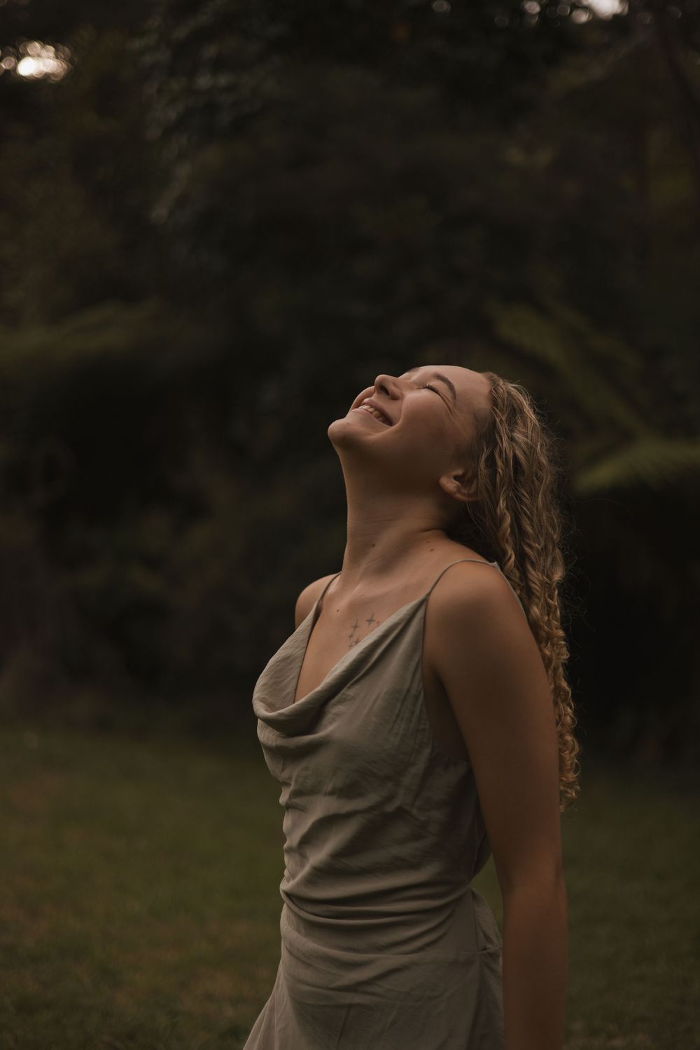 A joyful young woman with curly hair laughs with head tilted back, wearing a gray dress outdoors in a lush garden.