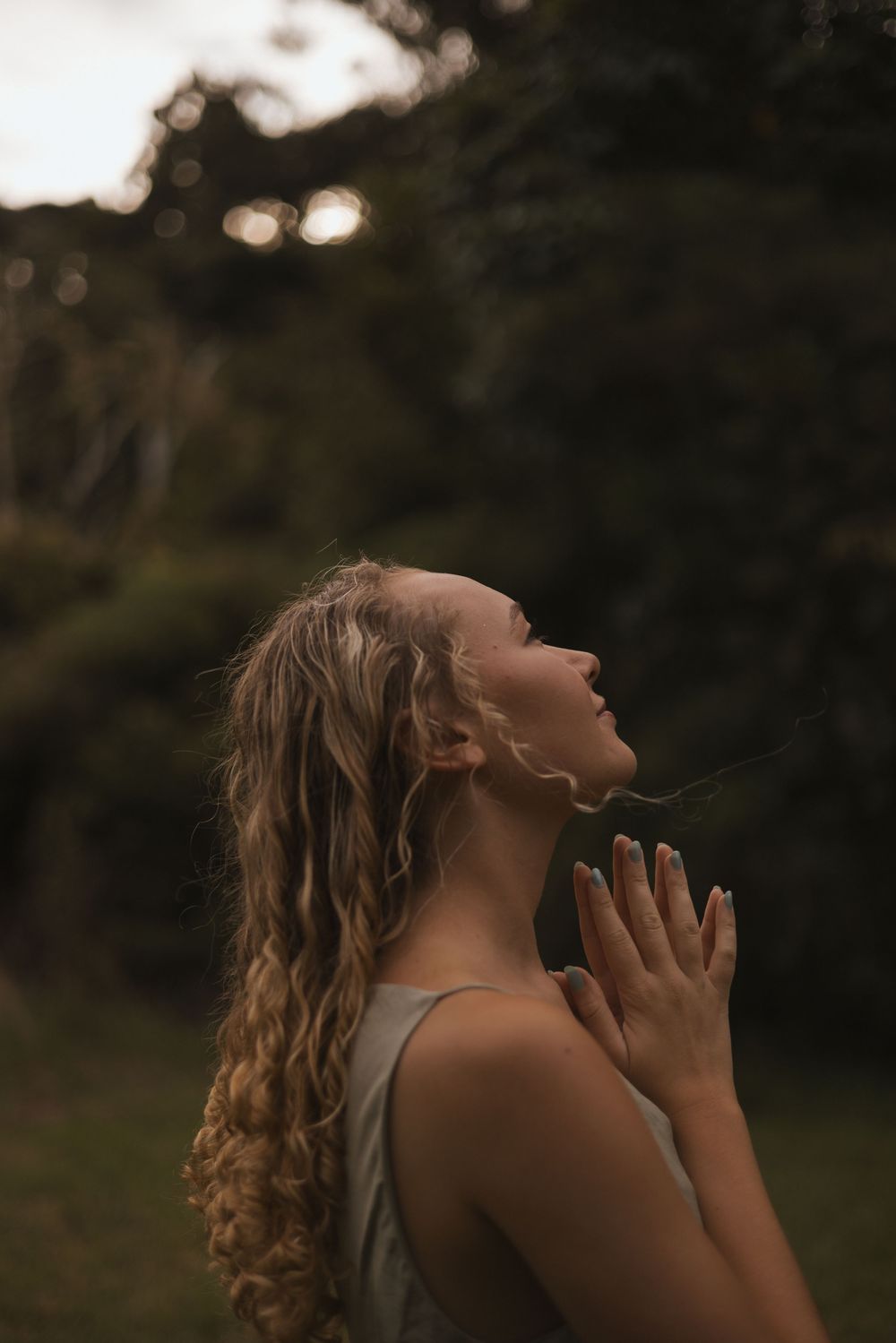 A young woman with curly blonde hair tilts her head back with hands in prayer pose outdoors at dusk.