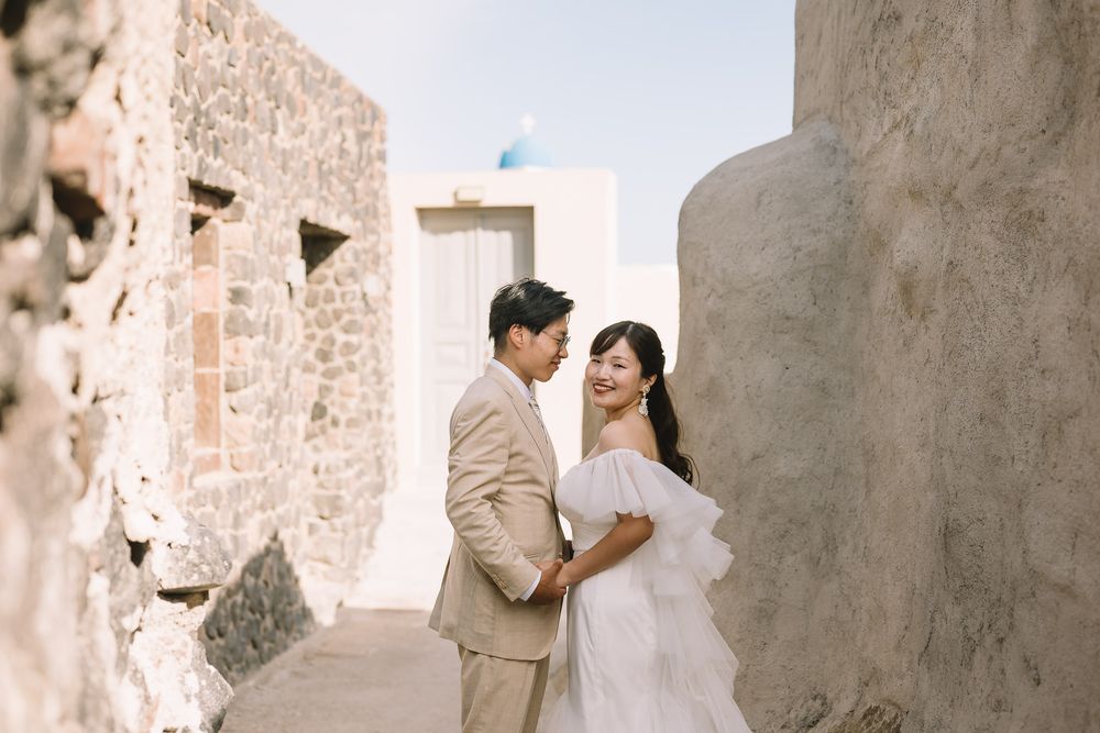 Couple in wedding attire poses romantically against stone walls in Santorini, Greece, with blue-domed church in background.