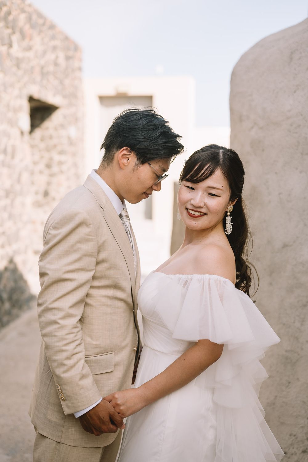Asian couple in wedding attire holding hands, groom in beige suit, bride in white off-shoulder dress, outdoor stone setting.
