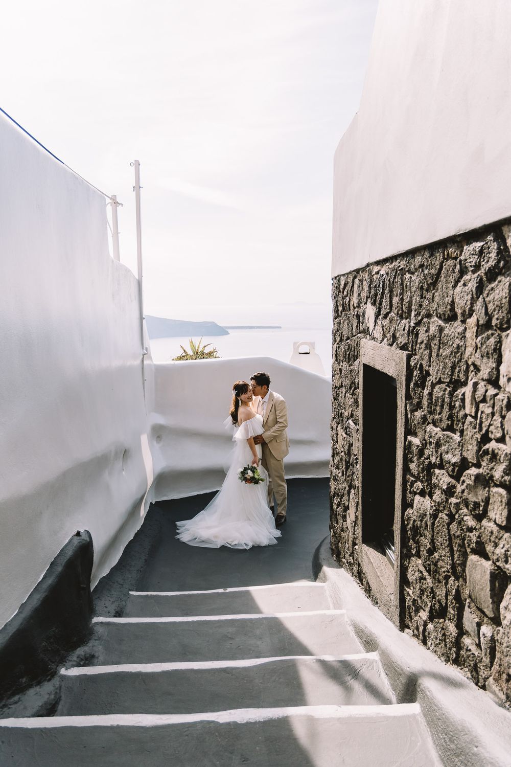 Bride and groom kissing on narrow Santorini steps with white-washed walls and caldera sea views.