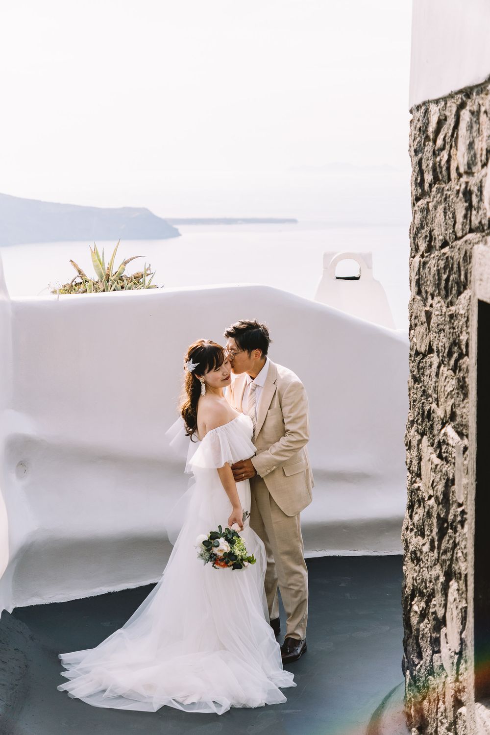Couple shares a romantic kiss in wedding attire with Santorini's white architecture and sea views backdrop.